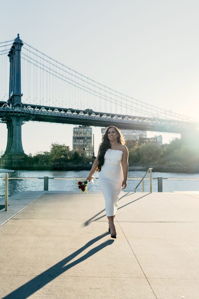 Woman in white dress walking near a bridge by the water.